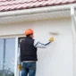 Worker wearing an orange safety helmet and gloves painting the exterior wall of a house with a roller brush near a glass door under a tiled roof
