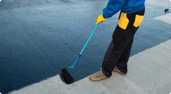 Worker applying black waterproof coating on a concrete surface using a paint roller with an extended handle