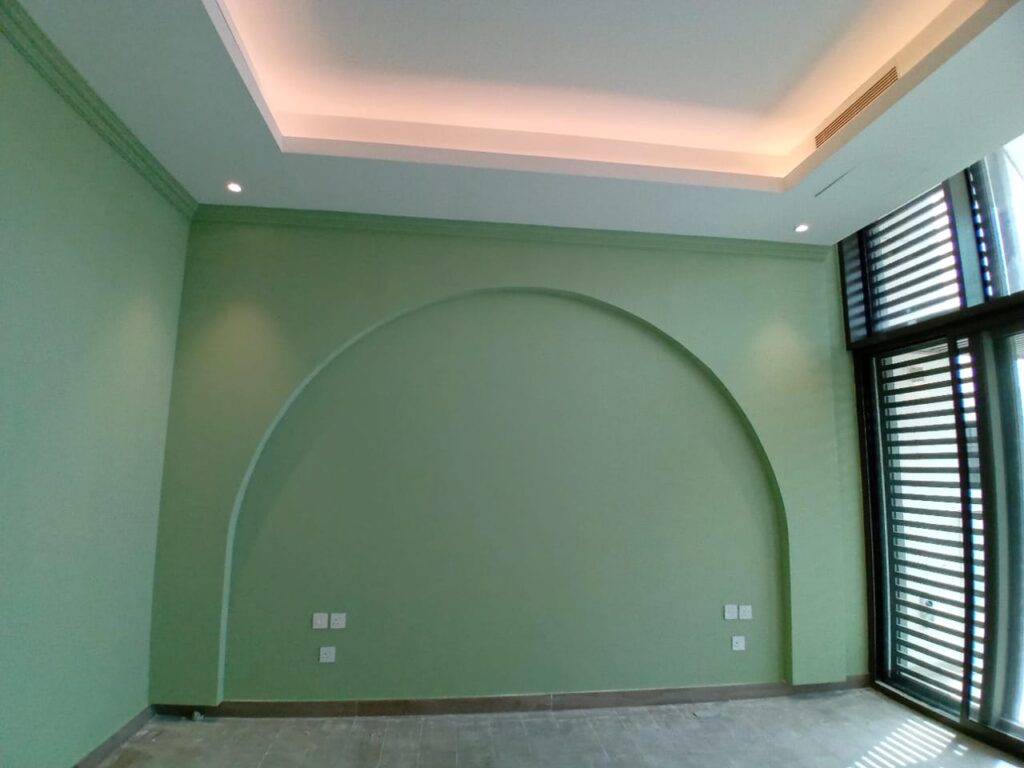 A modern empty room with light green walls, featuring a large arched design on one wall. The ceiling has recessed lighting with a soft glow, and there is a tall window with black vertical blinds allowing natural light to enter. The floor is tiled, and multiple electrical outlets are visible on the wall