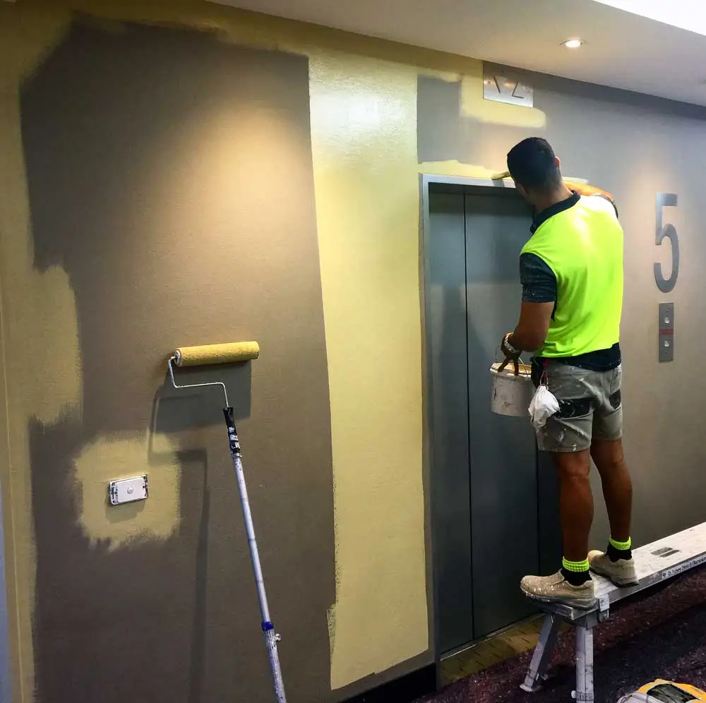 Painter standing on a small ladder painting the wall around an elevator door on the 5th floor, using a bucket and brush, with a paint roller on a pole nearby. The wall is partially painted from dark grey to light yellow